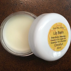 May include: A white jar with a yellow label that reads "Gusta Bliss Boutique Lip Balm Shea Butter, Beeswax, Coconut Oil, Argan Oil, Essential Oil if Scented Coconut .50 oz"