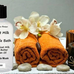 May include: A clear plastic bottle with a white label that reads "Goat Milk Bubble Bath" with ingredients listed as "With Goat Milk Shea Butter Argan Oil" and "Vanilla Musk". The bottle is 8 oz and is sitting on a white surface with orange towels, white flowers, and brown candles.