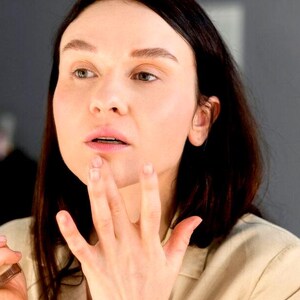 May include: A woman with long dark hair is applying makeup to her lips with her fingers. She is wearing a light beige shirt.