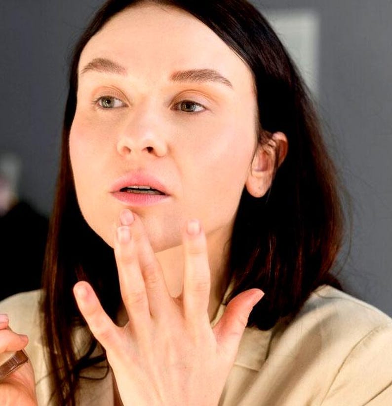 May include: A woman with long dark hair is applying makeup to her lips with her fingers. She is wearing a light beige shirt.
