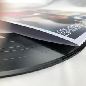 Puede incluir: Un disco de vinilo negro con surcos visibles, parcialmente cubierto por una funda de álbum blanca y negra. La funda del álbum presenta la palabra "Zenz" en letras blancas estilizadas. La imagen está tomada desde un ángulo bajo, resaltando la textura del disco.