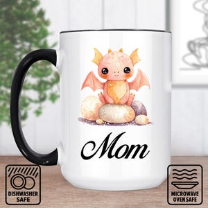 Pode incluir: Caneca de cerâmica branca com alça e borda pretas. Apresenta uma ilustração em aquarela de um dragão laranja fofo em rochas, com a palavra "Mom" em escrita preta. Adequado para máquina de lavar louça e micro-ondas.
