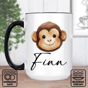 May include: White ceramic mug with a black handle and rim. The mug features a watercolor illustration of a monkey face with the name "Finn" in a script font. Dishwasher and microwave safe icons are also visible.