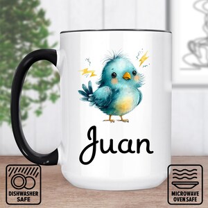 Peut inclure: Mug en céramique blanche avec une anse et un bord noirs. La tasse présente une illustration d'oiseau bleu avec le nom "Juan" en écriture noire. La tasse va au lave-vaisselle et au micro-ondes.