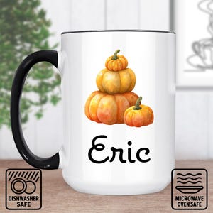 Könnte beinhalten: Weißer Keramikbecher mit schwarzem Henkel und Rand. Er zeigt eine Aquarellillustration von gestapelten Kürbissen in Orange- und Gelbtönen. Der Name "Eric" ist in schwarzer Schrift gedruckt. Spülmaschinen- und mikrowellengeeignete Symbole.