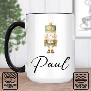 May include: White ceramic mug with a black handle and rim. The mug features a watercolor illustration of a nutcracker soldier and the name "Paul" in elegant script. Dishwasher and microwave safe icons are also visible.