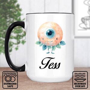 May include: White ceramic mug with a black handle and rim. The mug features a whimsical illustration of an eye with a smiling face, blue iris, and leafy accents. The name "Tess" is written below the illustration. Dishwasher and microwave safe.
