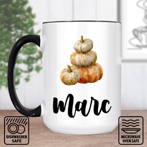 Könnte beinhalten: Weißer Keramikbecher mit schwarzem Henkel und Rand. Der Becher zeigt eine Aquarellillustration von gestapelten Kürbissen und den Namen "Marc" in schwarzer Schrift. Spülmaschinen- und mikrowellengeeignete Symbole sind ebenfalls sichtbar.
