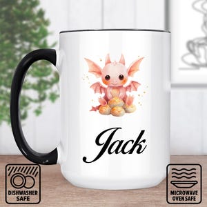 Pode incluir: Caneca de cerâmica branca com alça e borda pretas. A caneca apresenta uma ilustração em aquarela de um dragão rosa fofo com o nome "Jack" em escrita preta. Ícones de máquina de lavar louça e micro-ondas visíveis.