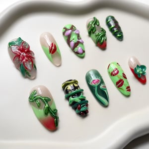 Pode incluir: Uma coleção de unhas artificiais com vários designs, incluindo motivos florais, de videira e de insetos. As unhas apresentam uma gama de cores, como vermelho, verde, rosa e branco, e estão dispostas numa bandeja branca.