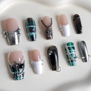 May include: A selection of false nails in diverse designs. Some nails are nude with silver details, others feature black and green plaid patterns, and some have white tips. Several nails are decorated with chains and charms.