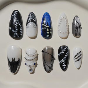 May include: Ten artificial nails with diverse designs. The nails showcase black, white, blue, and silver hues. Designs include floral patterns, cityscapes, and abstract art, with some adorned with pearls and gems. A variety of nail art.