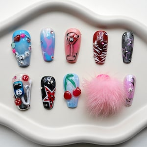 May include: A collection of ten artificial nails with various designs, including a martini glass, cherries, and stars. The nails are arranged on a white, cloud-shaped tray, with a pink fluffy accessory in the center.