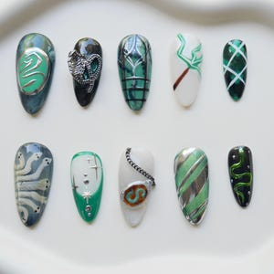 Puede incluir: Diez uñas postizas en forma de almendra, pintadas con diseños variados. El arte de uñas presenta rayas verdes y plateadas, un dragón plateado y patrones abstractos en verde, blanco y negro. Dispuestas sobre una bandeja ovalada blanca.