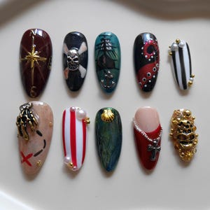 Puede incluir: Un juego de diez uñas postizas con temática pirata y náutica. Los diseños incluyen una brújula, una calavera, un barco, tentáculos de pulpo, rayas, un mapa del tesoro, una concha, una cruz y adornos dorados sobre las uñas individuales.