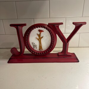 May include: A red metal JOY sign with a distressed finish. The letter O features a winter scene with a bird, tree, and small animal. The sign is designed to stand on a flat surface and is suitable for holiday decor.