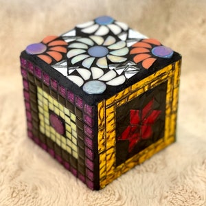 May include: A small, cube-shaped mosaic box with a black base and colorful glass tiles. The top of the box features a white and blue floral design, while the sides feature a yellow, red, and purple geometric pattern.