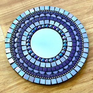 May include: A round mirror with a purple mosaic tile design. The mirror is surrounded by a border of iridescent tiles and small silver beads.