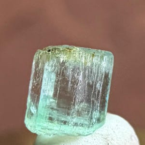 May include: A translucent, pale green, rectangular crystal specimen with vertical striations along its sides. The mineral features a clear, glassy texture with subtle internal fractures and a slightly yellowish tint near the top edge.