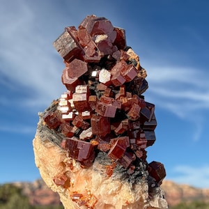 May include: A striking mineral specimen featuring a cluster of deep red, cubic crystals atop a light-colored matrix. The crystals exhibit a rich, glossy appearance, contrasting with the rougher texture of the base. The background shows a clear blue sky and distant landscape.