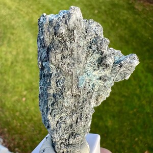 May include: A rough, textured mineral specimen with a mix of gray and teal hues. The stone has a jagged, irregular shape, with a crystalline structure visible on the surface. The mineral is held on a white base, with a blurred green background.