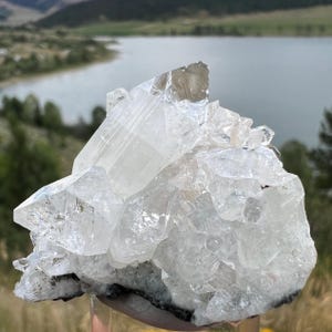 May include: A cluster of clear quartz crystals with multiple points and facets. The crystals are translucent, with a slightly cloudy appearance in some areas. The quartz sits on a dark base, with a natural landscape in the background.