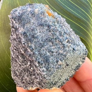 May include: A close-up of a mineral specimen with a textured surface. The rock has a mix of blue, gray, and orange hues. The surface is covered in small, crystalline structures, giving it a fuzzy appearance. The mineral is set against a green leaf.