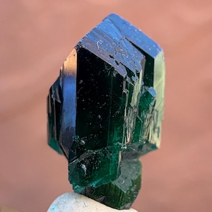 May include: A dark green crystal with a faceted, geometric shape. The crystal has a glossy surface and appears to be translucent, with light reflecting off its edges. The crystal is mounted on a small, light-colored base, with a blurred background.