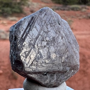 May include: A large, dark gray, faceted crystal with striations and a rough, natural texture. The crystal is sitting on a small, gray rock.