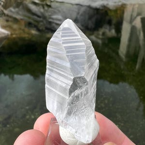 May include: A clear, faceted crystal point with striations, displayed on a small white base. The crystal is a light, translucent white and has a pointed tip.