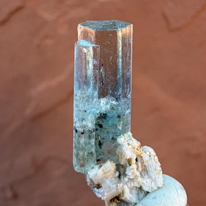 May include: A translucent, hexagonal-shaped aquamarine crystal with a smaller, parallel crystal formation. The main crystal is a light blue color, with some darker inclusions. The base features a cluster of white and beige mineral formations.