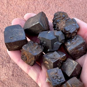 May include: A collection of dark, geometric mineral specimens, held in a hand. The stones are primarily dark brown and black, with a rough, textured surface. The shapes vary, with some appearing more cubic and others with irregular facets.