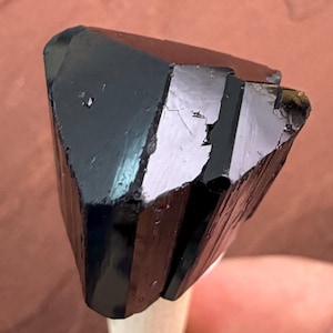 May include: A close-up of a black tourmaline crystal. The crystal is a deep, glossy black with a faceted, geometric shape. The surface reflects light, showing subtle variations in color and texture. The crystal is held on a wooden dowel.