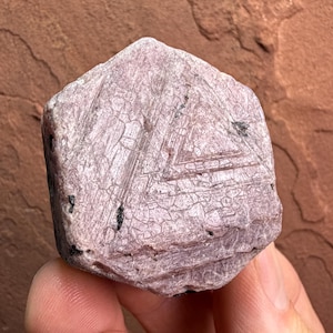 May include: A hexagonal raw corundum crystal with a dusty mauve and light pink coloration. The surface displays a distinct geometric triangular pattern with etched lines and small black mineral inclusions embedded within the crystalline structure.