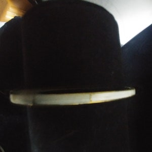 May include: A black hat with a white brim. The hat is set against a dark background, with a bright light source illuminating the top of the hat. The brim has a slight yellowing.