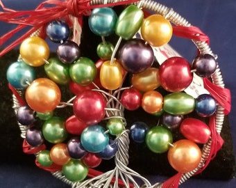 May include: A handcrafted, wire-wrapped tree of life ornament. The tree features a silver wire trunk and branches, adorned with colorful, iridescent beads in shades of red, orange, yellow, green, blue, and purple. The ornament is encircled with red ribbon.