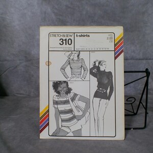 May include: A white paper sewing pattern envelope for Stretch & Sew t-shirts number 310. The front features black line drawings of three t-shirt styles, text including bust sizes 28-44, and diagonal multi-coloured stripes on the left and right edges.