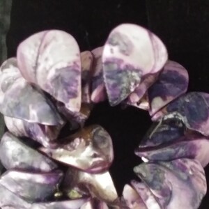 Puede incluir: Un brazalete hecho de conchas iridiscentes de color púrpura. Las conchas están dispuestas en un patrón circular, creando un accesorio único y llamativo. El brazalete está sobre un fondo oscuro.