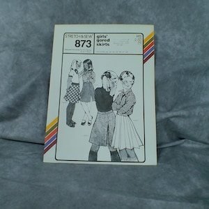May include: A vintage Stretch & Sew 873 sewing pattern envelope for girls' gored skirts. The white paper cover features black line illustrations of four different skirt styles, with text including the pattern number 873 and size range 22-28.