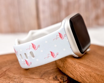 May include: A white Apple Watch band with a light blue background and pink flamingo print. The band has white polka dots and is made of silicone.