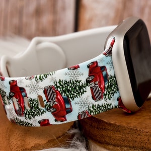 Op de afbeelding: Witte Apple Watch band met een kerstthema print met rode pick-up trucks, groene dennenbomen en sneeuwvlokken op een lichtblauwe achtergrond.