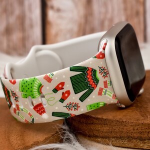 Op de afbeelding: Een wit Apple Watch bandje met een kerstthema met groene en rode truien, wanten en hulsttakken.