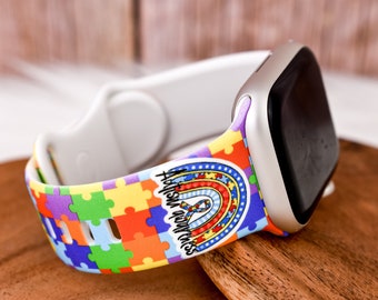 May include: A white Apple Watch band with a colorful puzzle piece design and the text "Autism Awareness" on a rainbow background.