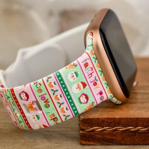 Könnte beinhalten: Eine weiße Apple Watch mit einem pink- und grünfarbenen Weihnachtsband. Das Band zeigt ein sich wiederholendes Muster aus Schneeflocken, Rentieren, Weihnachtsmann und den Worten "Frieden, Liebe, Hoffnung, Glaube, Freude".