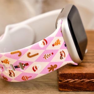 Könnte beinhalten: Ein rosafarbenes und weißes Apple Watch Armband mit Weihnachtsmotiv. Das Armband zeigt ein sich wiederholendes Muster aus Zuckerstangen, Lebkuchenmännern, Weihnachtsbäumen und Weihnachtsmann.