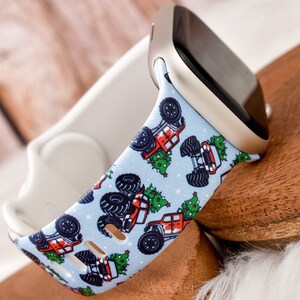 Könnte beinhalten: Ein blaues und weißes Apple Watch Armband mit einem Weihnachtsdesign, das rote Monstertrucks mit Weihnachtsbäumen zeigt. Das Armband ist aus Silikon und kompatibel mit Apple Watch Series 4 und höher.