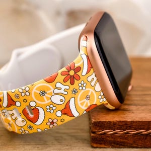 Könnte beinhalten: Ein gelbes und weißes Armband mit Mustern aus Peace-Zeichen, Blumen und Weihnachtsmützen. Das Armband ist an einer roségoldenen Apple Watch befestigt.