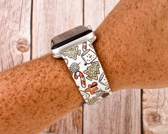May include: A white Apple Watch band with a Christmas-themed print featuring gingerbread men, candy canes, and Christmas trees. The band is on a person's wrist.