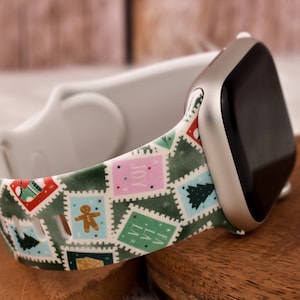 Op de afbeelding: Een witte Apple Watch met een groen, wit en roze kerstband. De band heeft een herhalend patroon van postzegels met verschillende kerstafbeeldingen, waaronder een peperkoekman, een kerstboom en het woord "Vreugde".