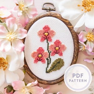 May include: A cross-stitch floral design featuring three pink flowers with yellow centers and green leaves, set in an oval wooden frame. The words "PDF PATTERN" are in a circle on the bottom right. Surrounded by pink and white flowers.